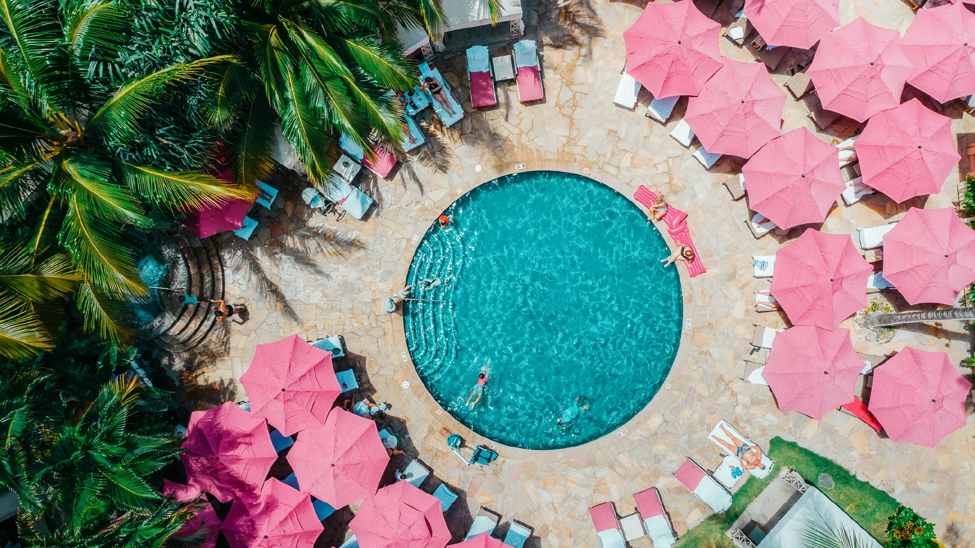 Aerial view of the Royal Hawaiian pool surrounded by pink umbrellas, palm trees, and turquoise water.