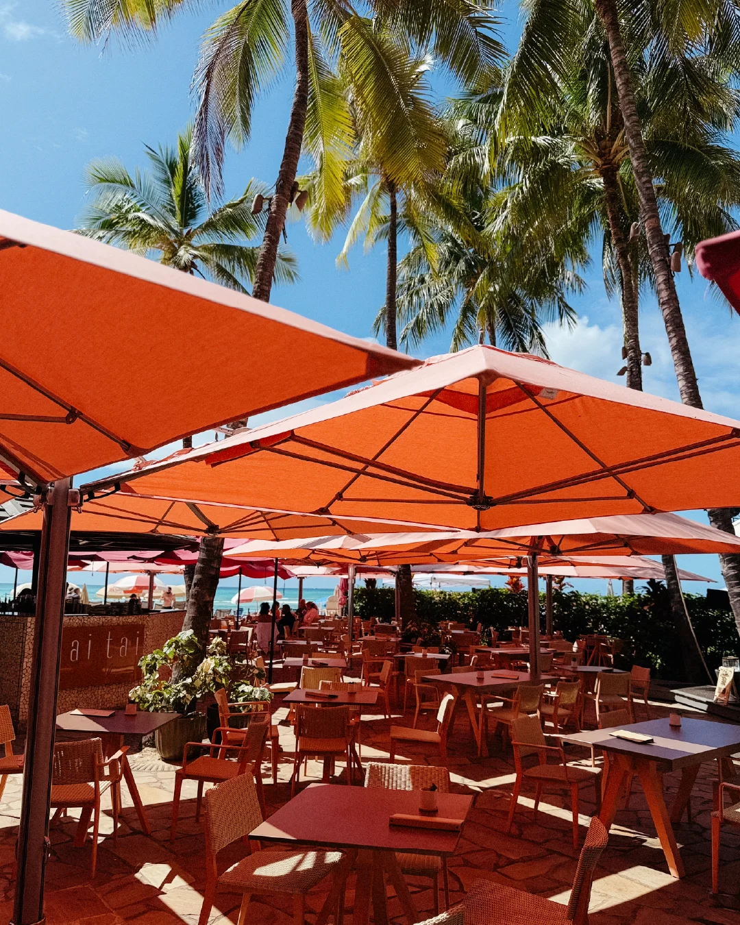 Outdoor dining area near the beach with coral umbrellas and palm trees at the Royal Hawaiian.