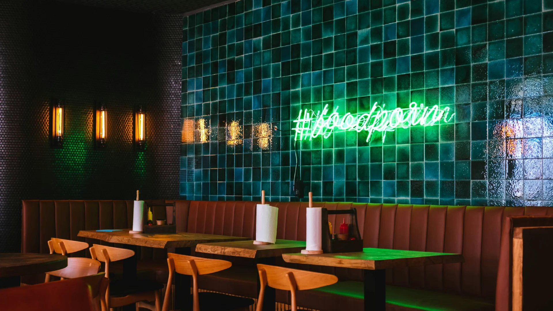 Wide view of a restaurant dining room with glossy green tile, booth seating, wood tables, and glowing neon wall signage.