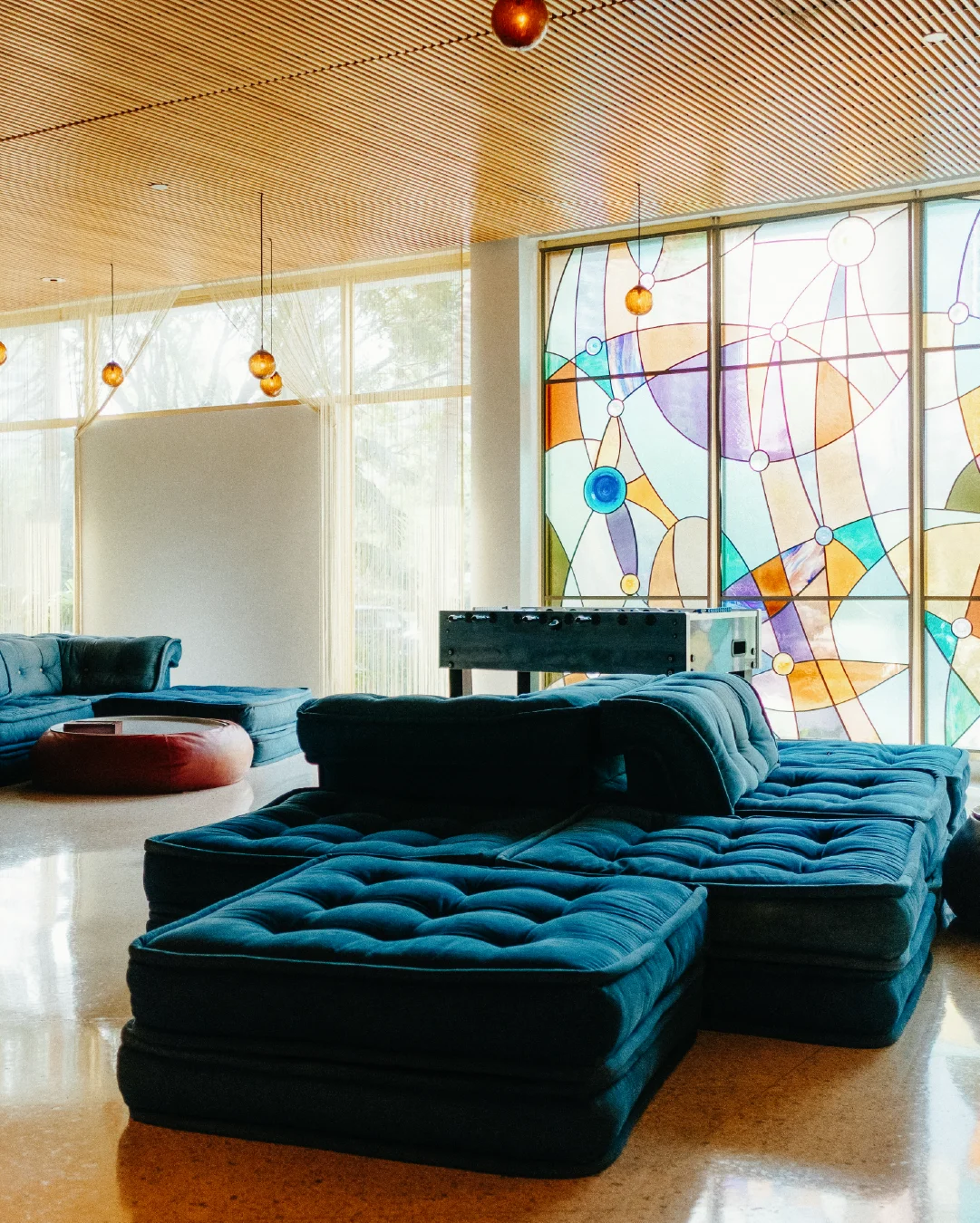 Lounge with deep blue modular seating, warm pendant lights, and colourful stained-glass-style windows.
