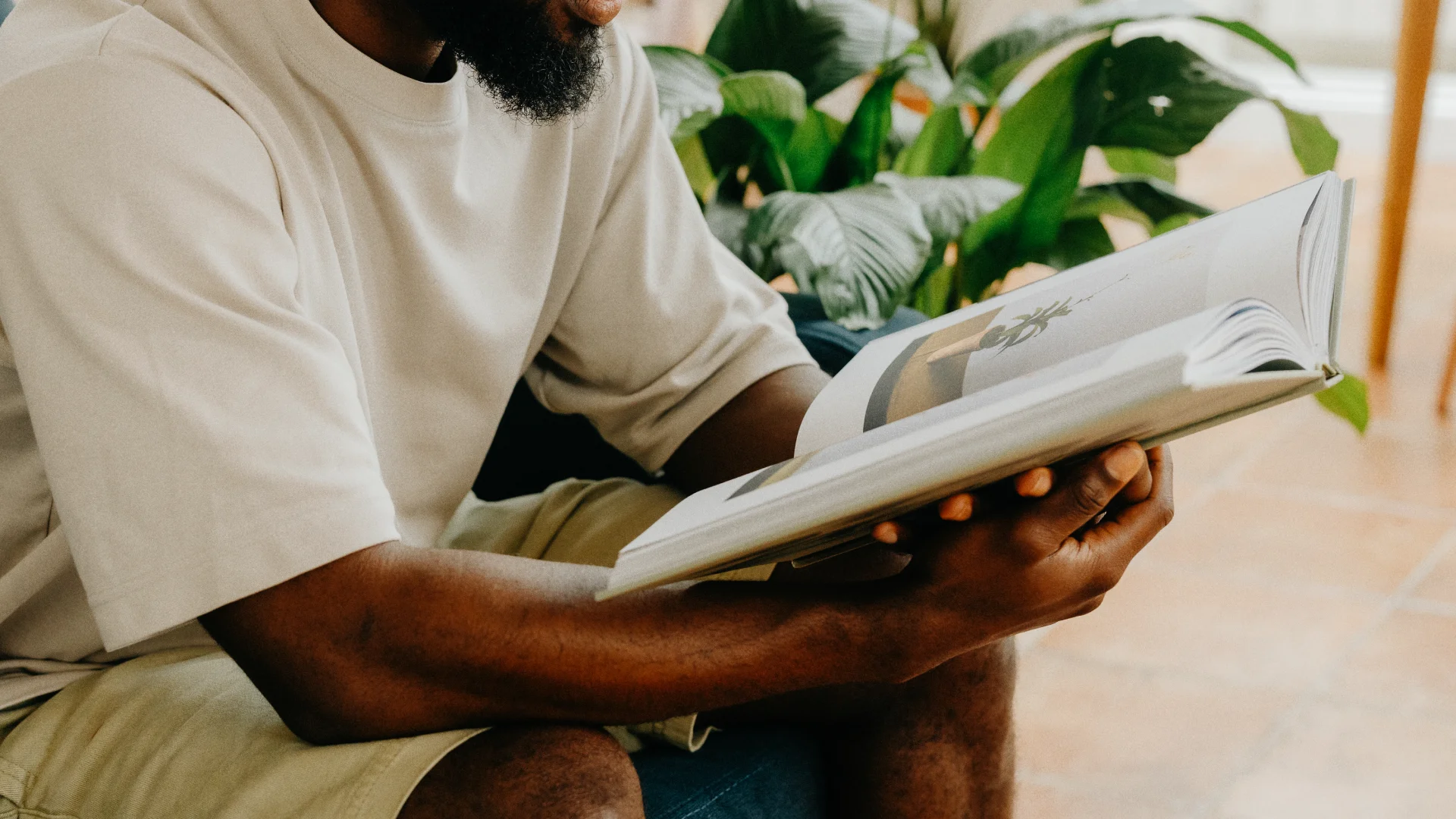 Hotel guest seated indoors reading a large printed book beside lush green plants in a calm hospitality setting.