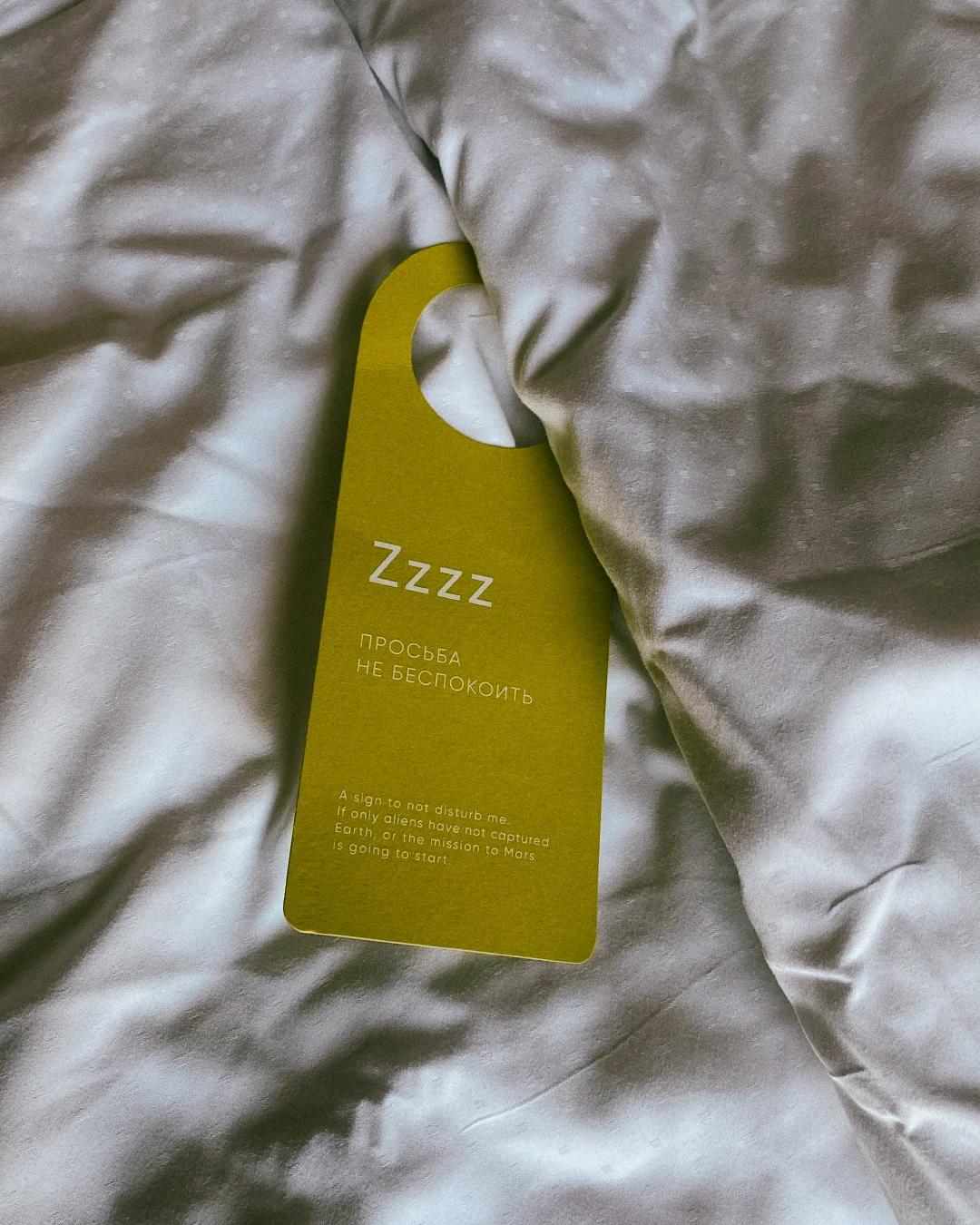 Green printed do not disturb door hanger resting on white hotel bedding in a guest room.