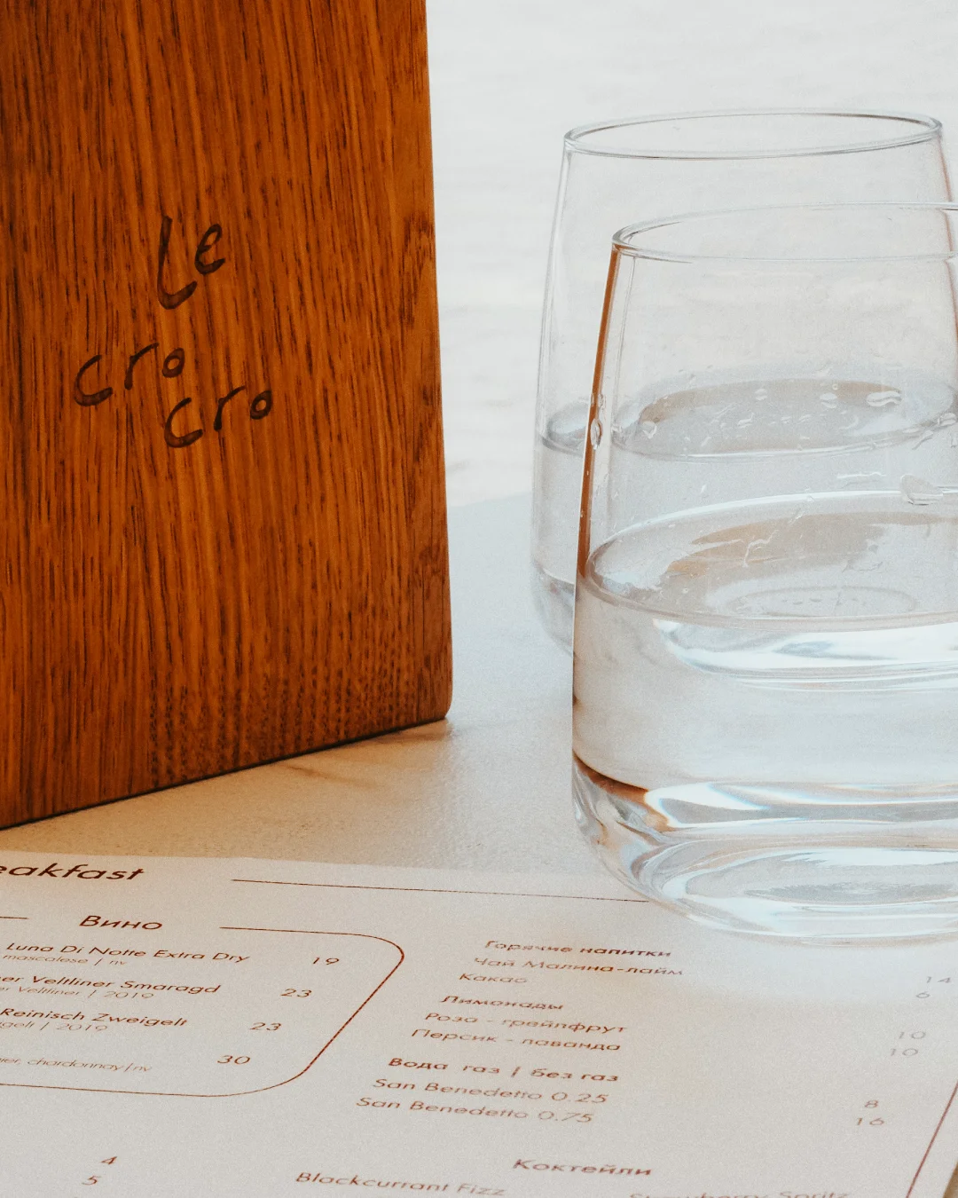Printed restaurant menu on a table beside water glasses and a wooden menu stand in a refined dining setting.
