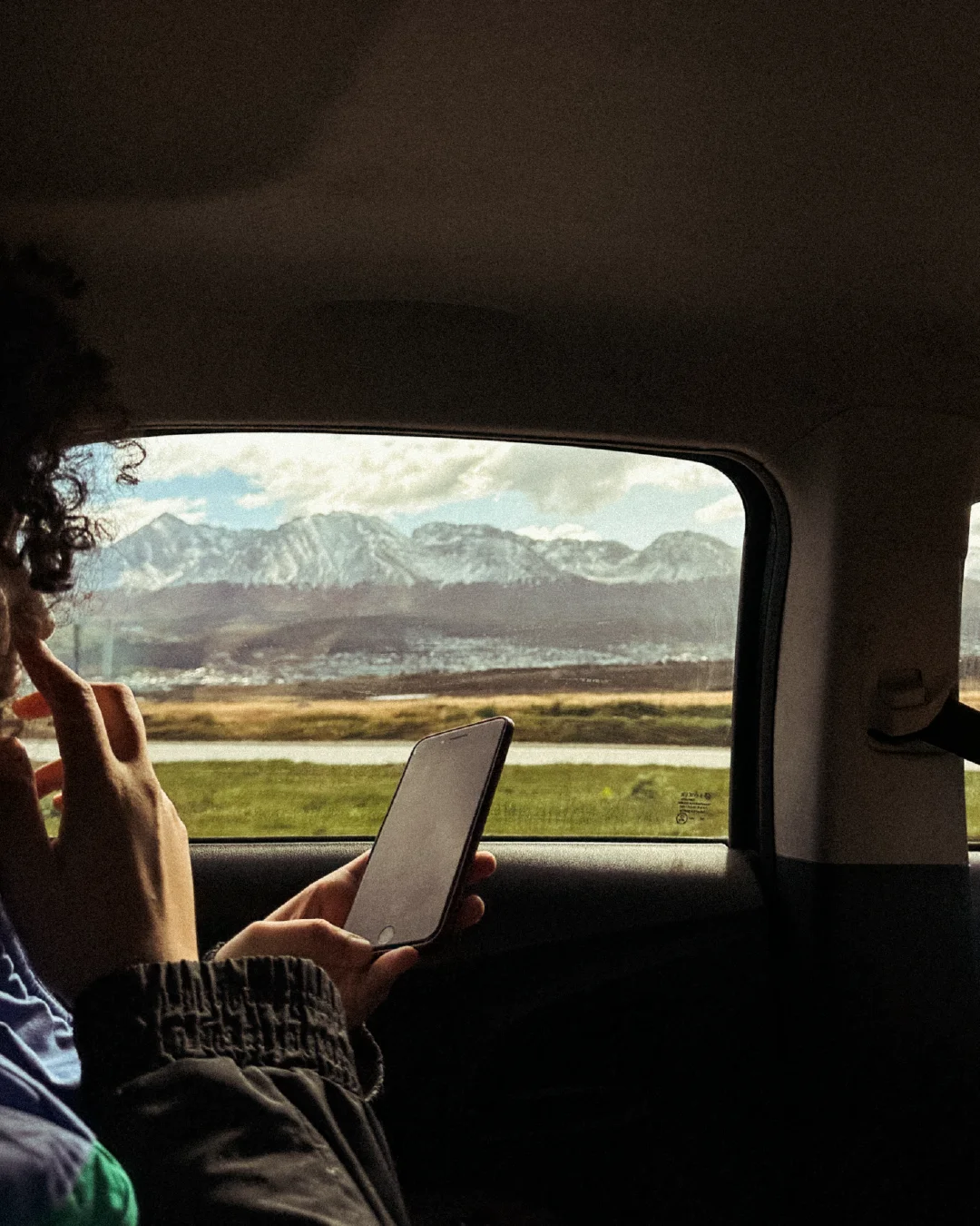 A traveller holds a phone in the back seat of a car while passing a mountain landscape.