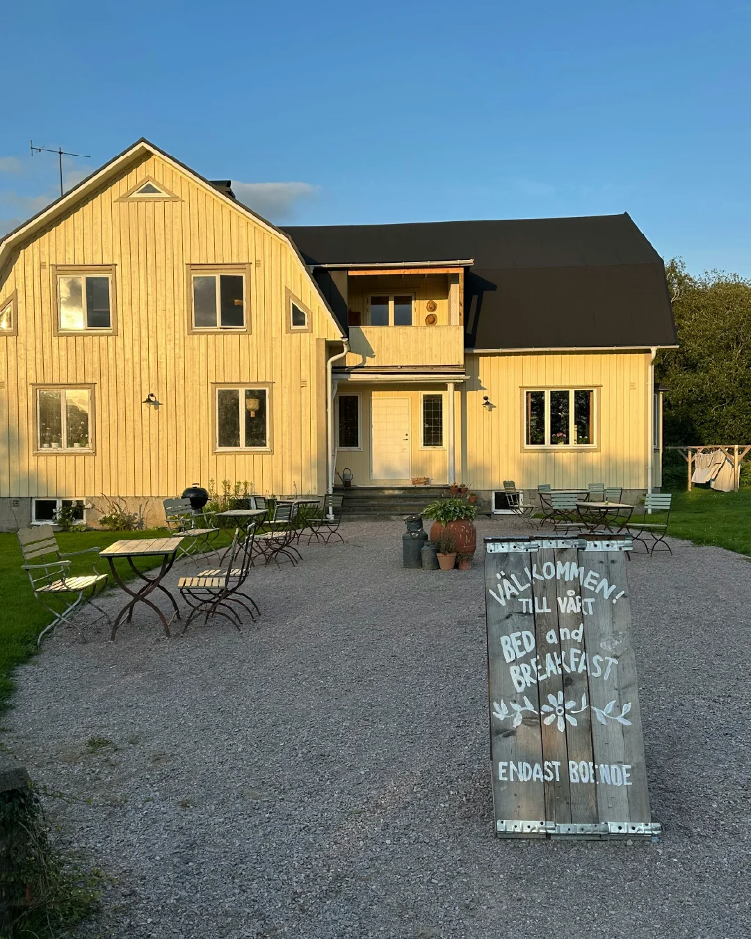 A hand-painted bed and breakfast welcome sign outside a yellow guesthouse in warm evening light.
