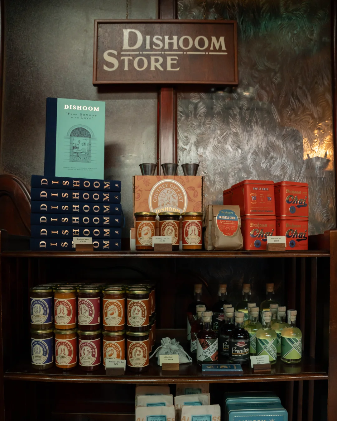 Dishoom Store display with branded cookbooks, chutneys, chai tins, jars, bottles, and gift products arranged on dark wooden shelves.