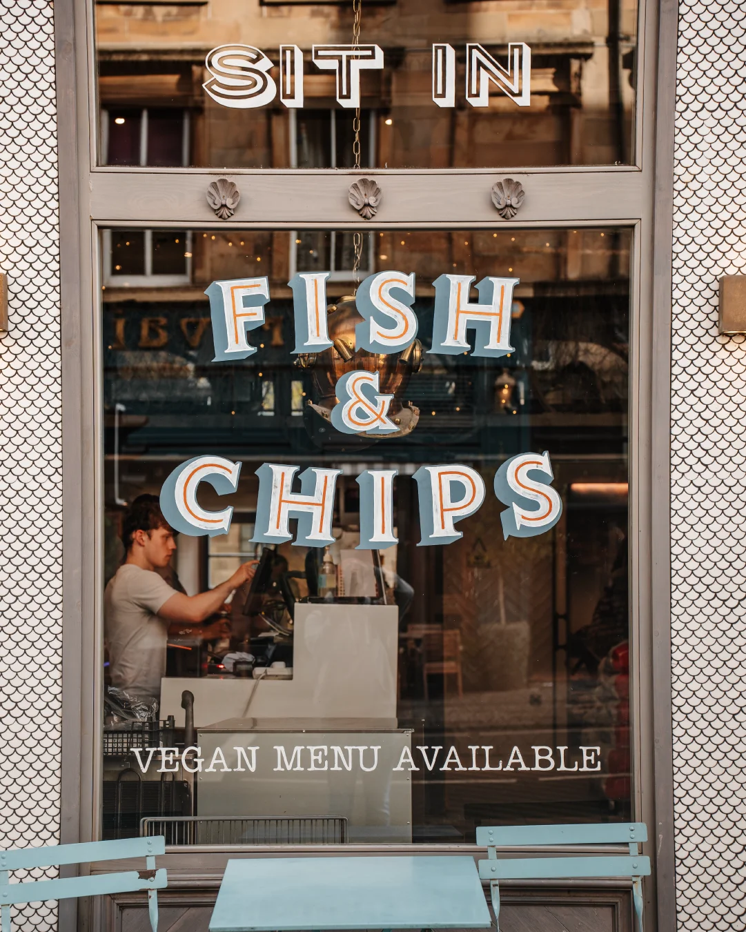 Restaurant window with large hand-painted “Fish & Chips” lettering, “Sit In” signage, and “Vegan Menu Available” text on the glass.