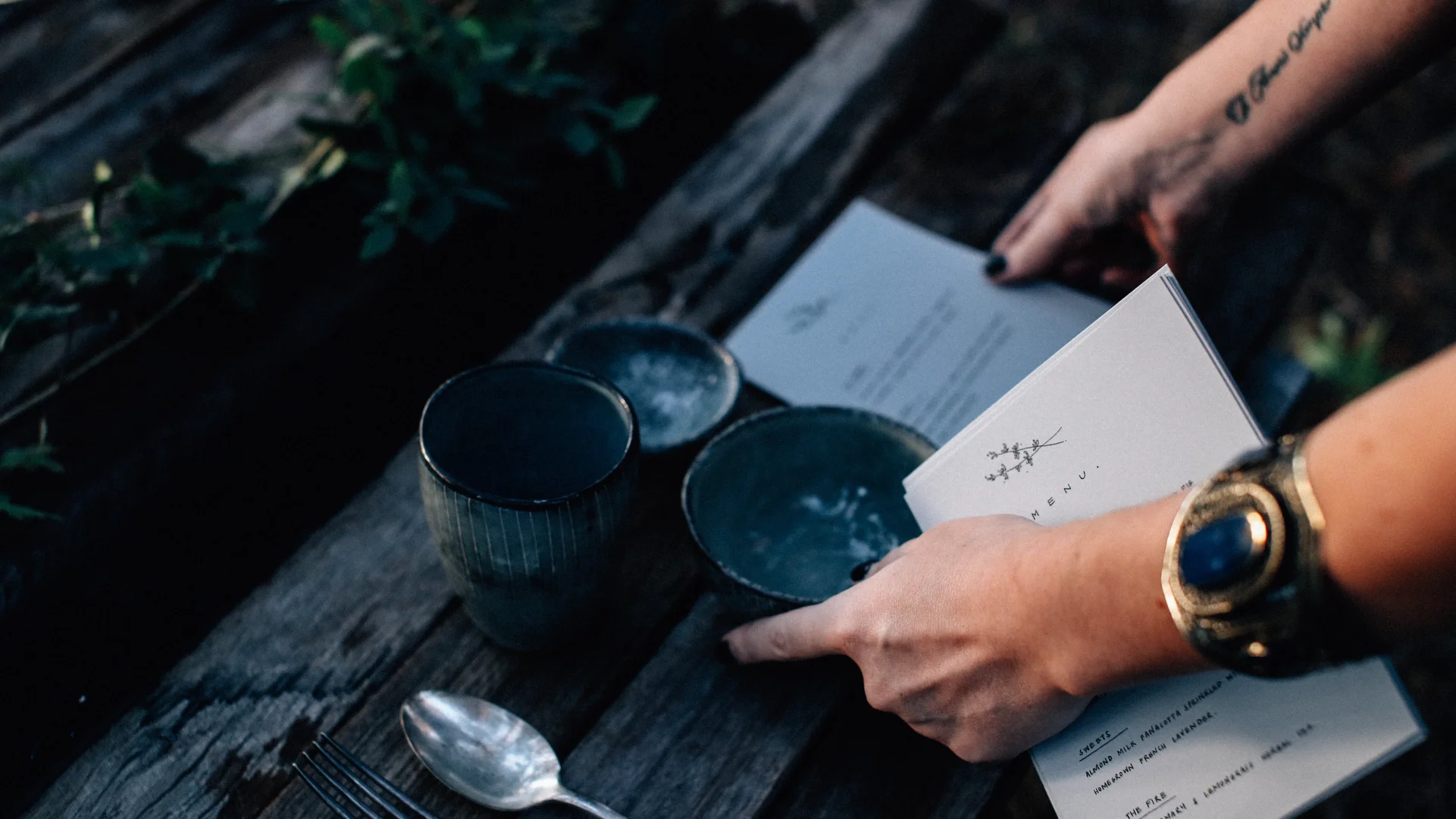 Close-up of a guest holding a printed restaurant menu beside ceramic cups, bowls, a spoon, and fork on a dark wooden table.