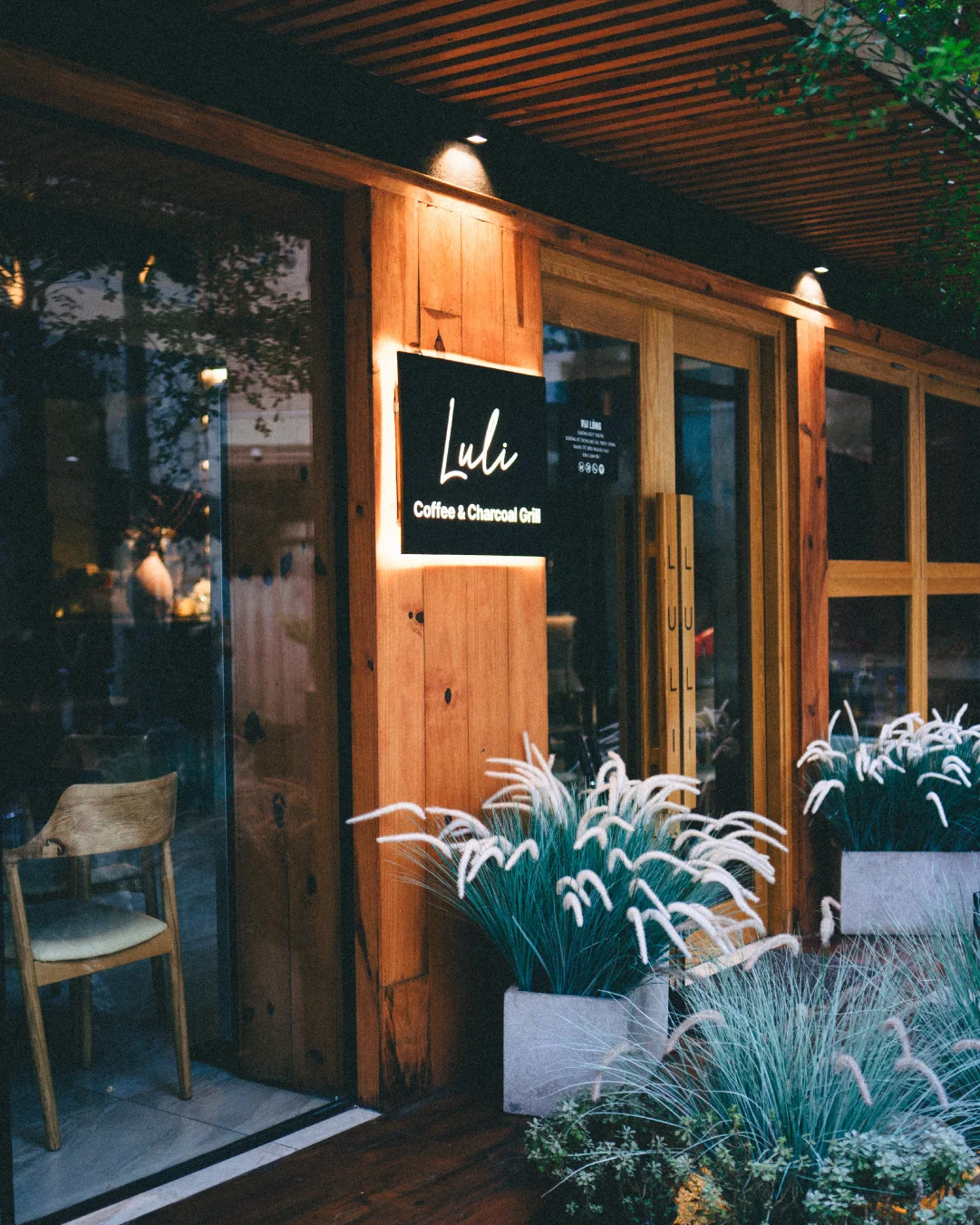 Evening exterior of Luli Coffee & Charcoal Grill with illuminated signage, warm wood cladding, large windows, and ornamental grasses by the entrance.