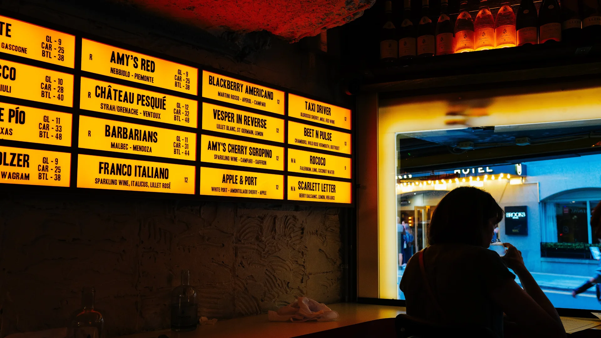 Interior restaurant bar with bright yellow illuminated menu boards listing wine and cocktail options above a seated guest silhouette.