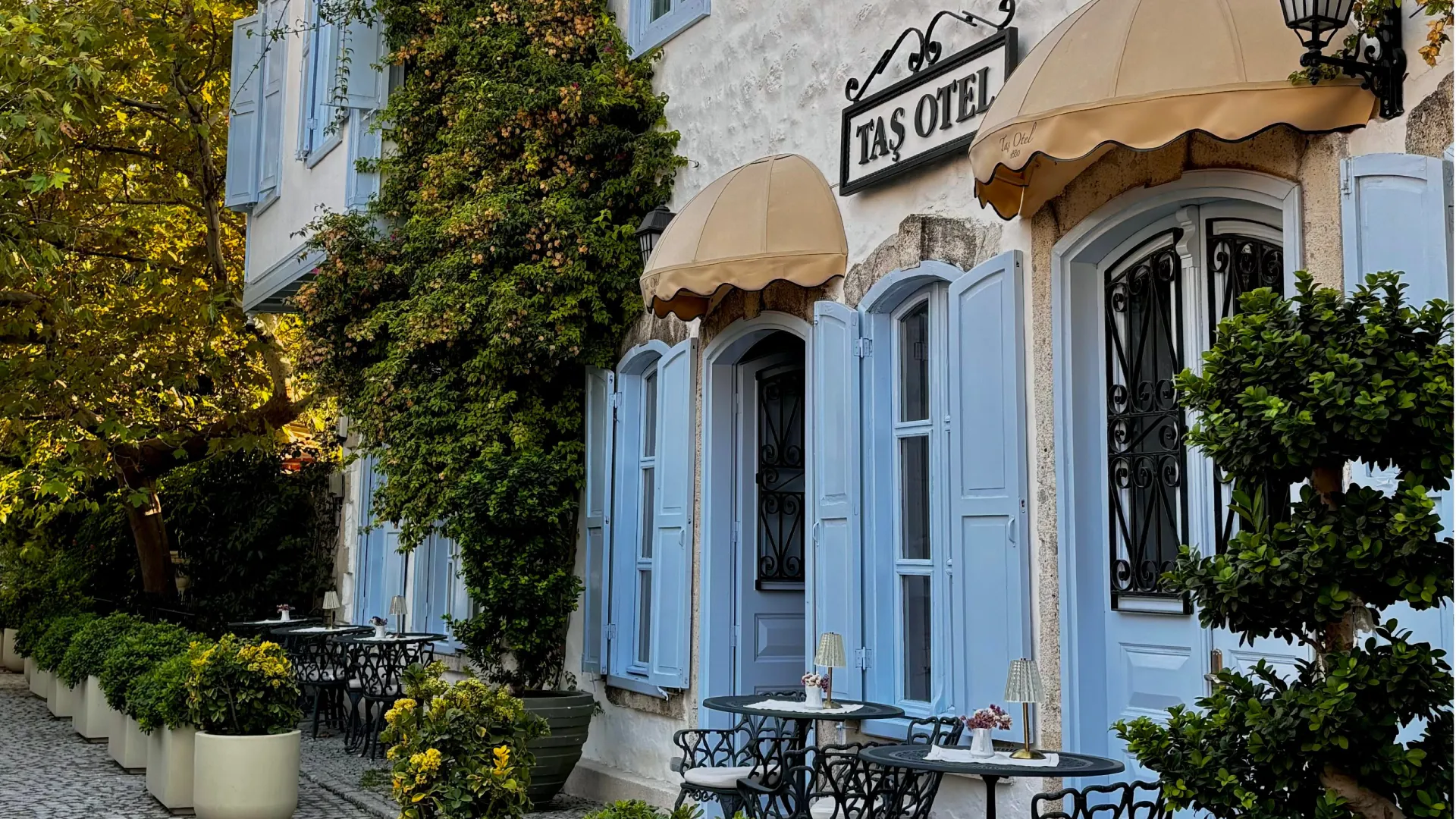 Exterior of a historic boutique hotel with pale blue shutters, cream awnings, greenery, and outdoor café tables.
