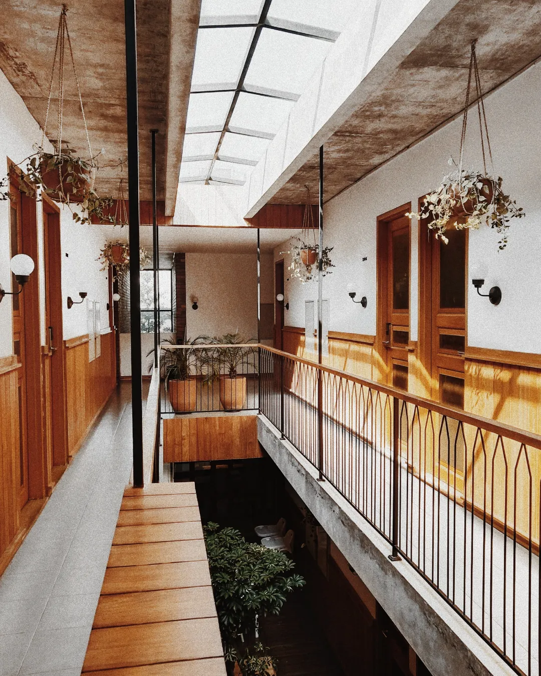 Boutique hotel hallway with warm wood doors, hanging plants, skylights, and black railings.