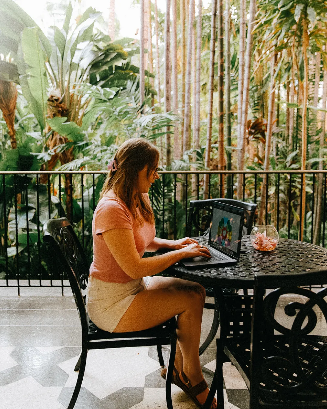 Guest working on a laptop at an outdoor table surrounded by tropical plants inside a hotel courtyard.