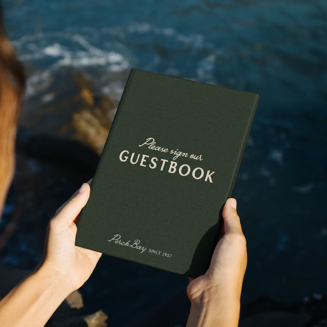 Person holding a dark green Perch Bay guestbook beside the rocky waterfront.