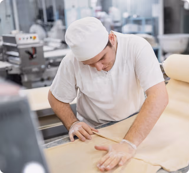 Man in white uniform and cap working with dough on a table in a commercial kitchen.