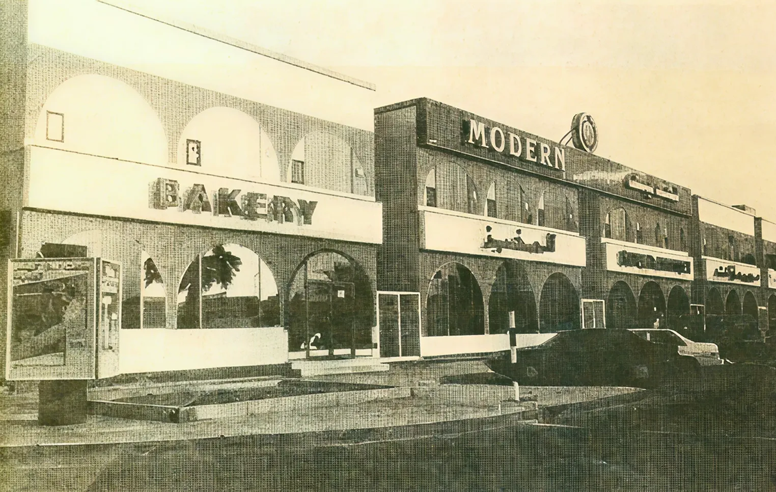 Vintage sepia-toned photo of a brick commercial building with arched windows and signage including 'MODERN' and 'BAKERY,' with cars parked in front.