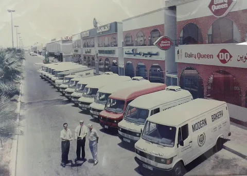 Row of bakery delivery vans parked near a Dairy Queen building with three men standing in the foreground on a sunny day.