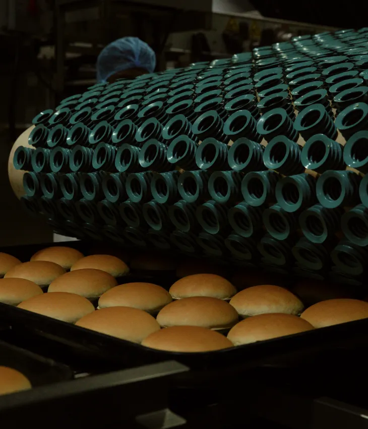 Rows of freshly baked hamburger buns on baking trays under a machine with multiple round suction cups in a bakery production line.