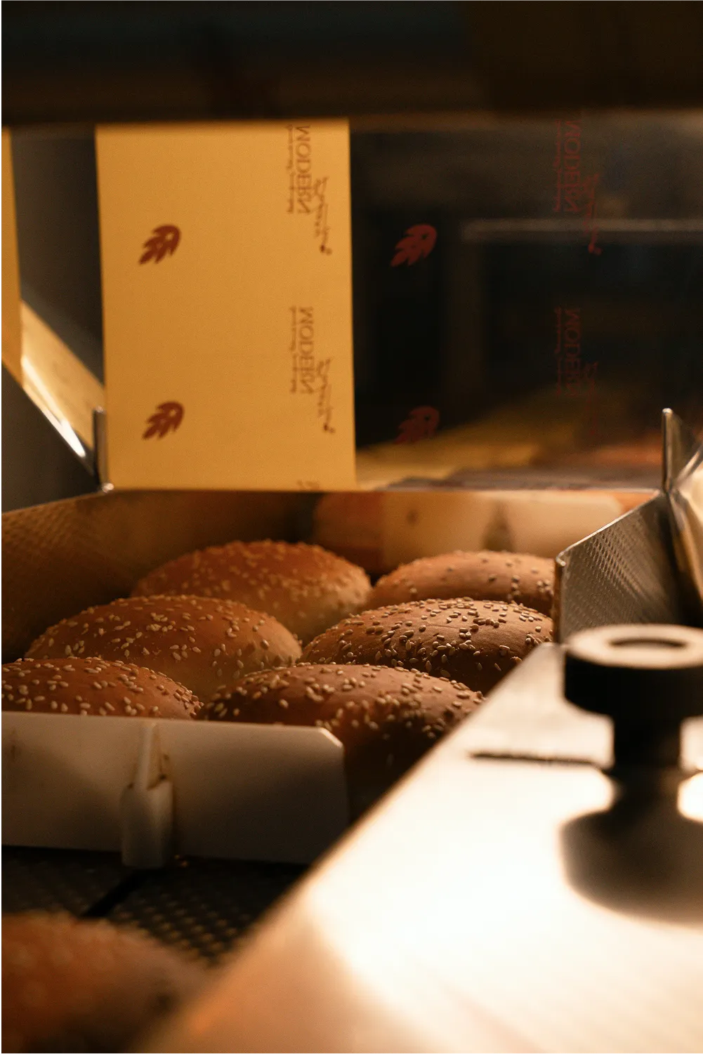 Several sesame seed hamburger buns inside a partially open cardboard box on a conveyor belt.