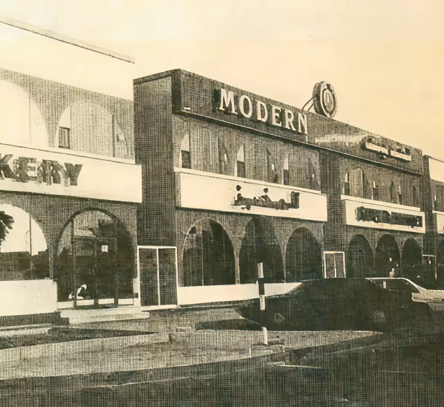 Vintage black and white photo of a brick commercial building with arched windows, a sign reading 'MODERN,' and a few parked cars.