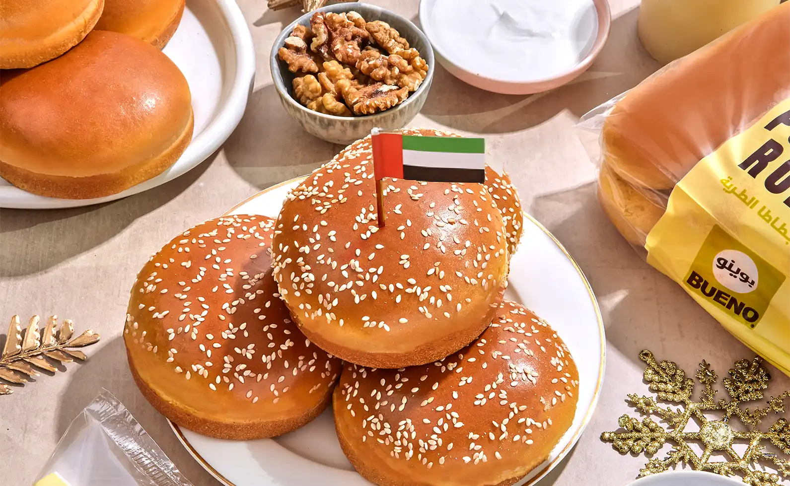 Plate with three sesame seed burger buns, one topped with a small United Arab Emirates flag, surrounded by bread rolls, walnuts in a bowl, and a yellow bread package labeled BUENO.