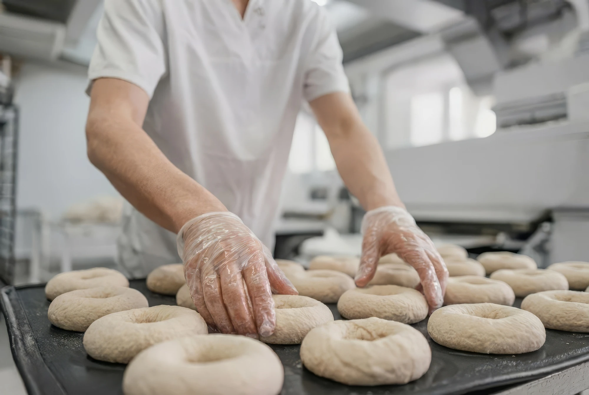 Person wearing plastic gloves shaping dough rings on a tray in an industrial bakery.