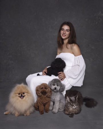 Giovane donna in abito bianco con cane barboncino nero in grembo, circondata da Pomerania, barboncino rosso, barboncino grigio e gatto grigio, ritratto in studio.