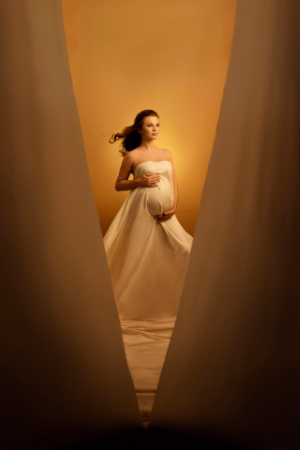 Donna incinta avvolta in tessuto chiaro tra tende dorate, atmosfera calda e luminosa, servizio fotografico di maternità a Bologna.