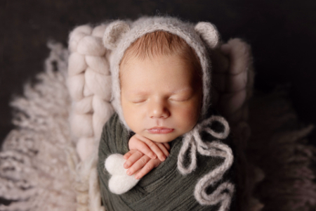 Neonato avvolto in fascia verde con cappellino orsetto e decorazione a cuore, mani sotto il mento, fotografia newborn delicata.