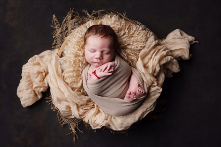 Neonato addormentato avvolto in tessuto beige, posato in cestino circolare con drappeggi crema, fotografia fine art in luce soffusa.