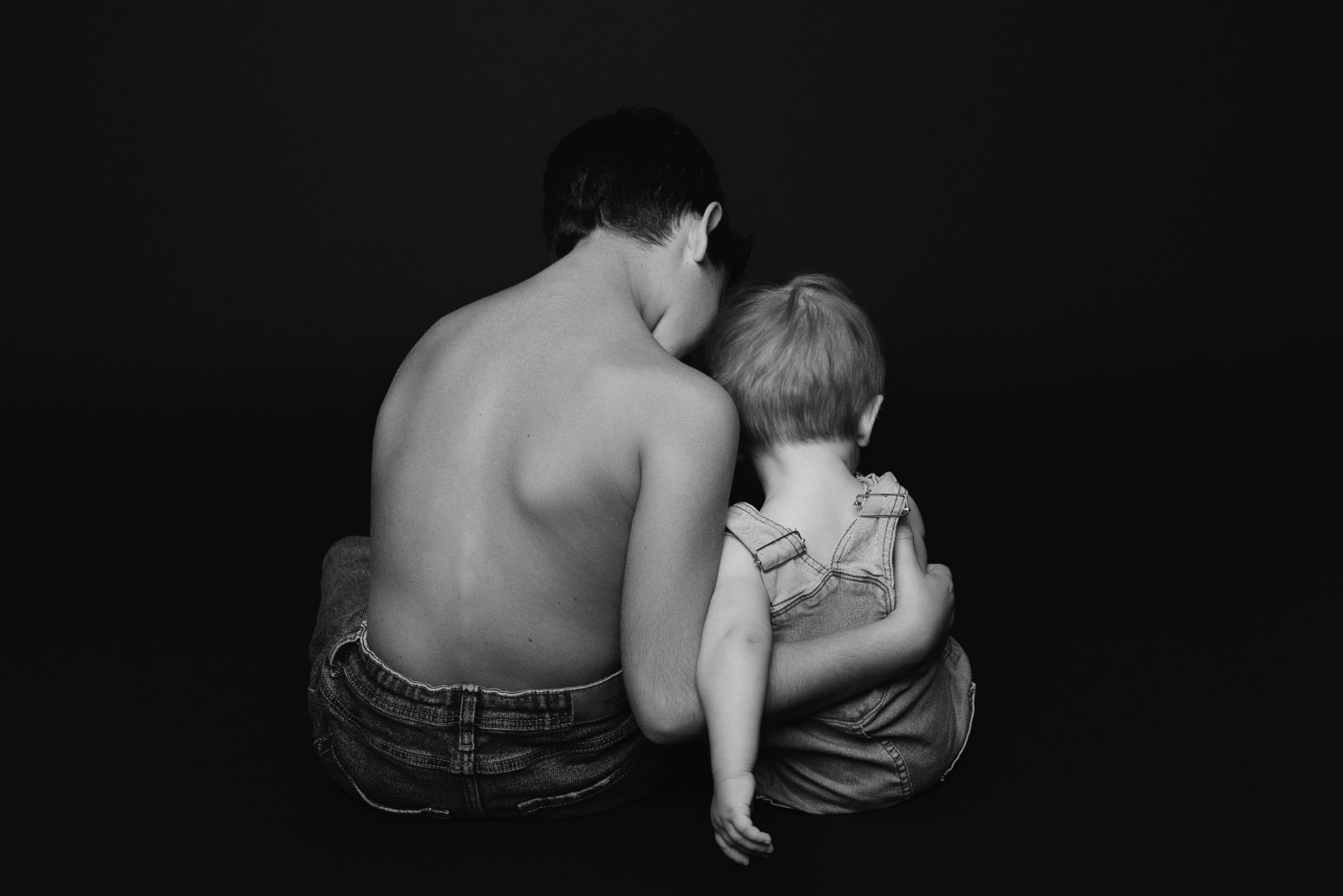 Due bambini seduti di spalle contro uno sfondo scuro. Il maggiore, a torso nudo, abbraccia il più piccolo che indossa una salopette. L’immagine evoca affetto, protezione e legame familiare.