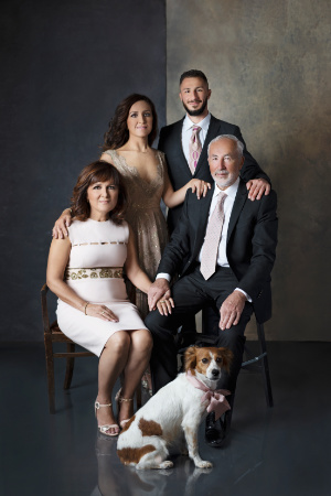 Servizio fotografico di famiglia con cane a Bologna – Claudia Spaziani – Due adulti, due giovani e cane con fiocco rosa, abbigliamento elegante, ritratto in studio con sfondo sfumato.