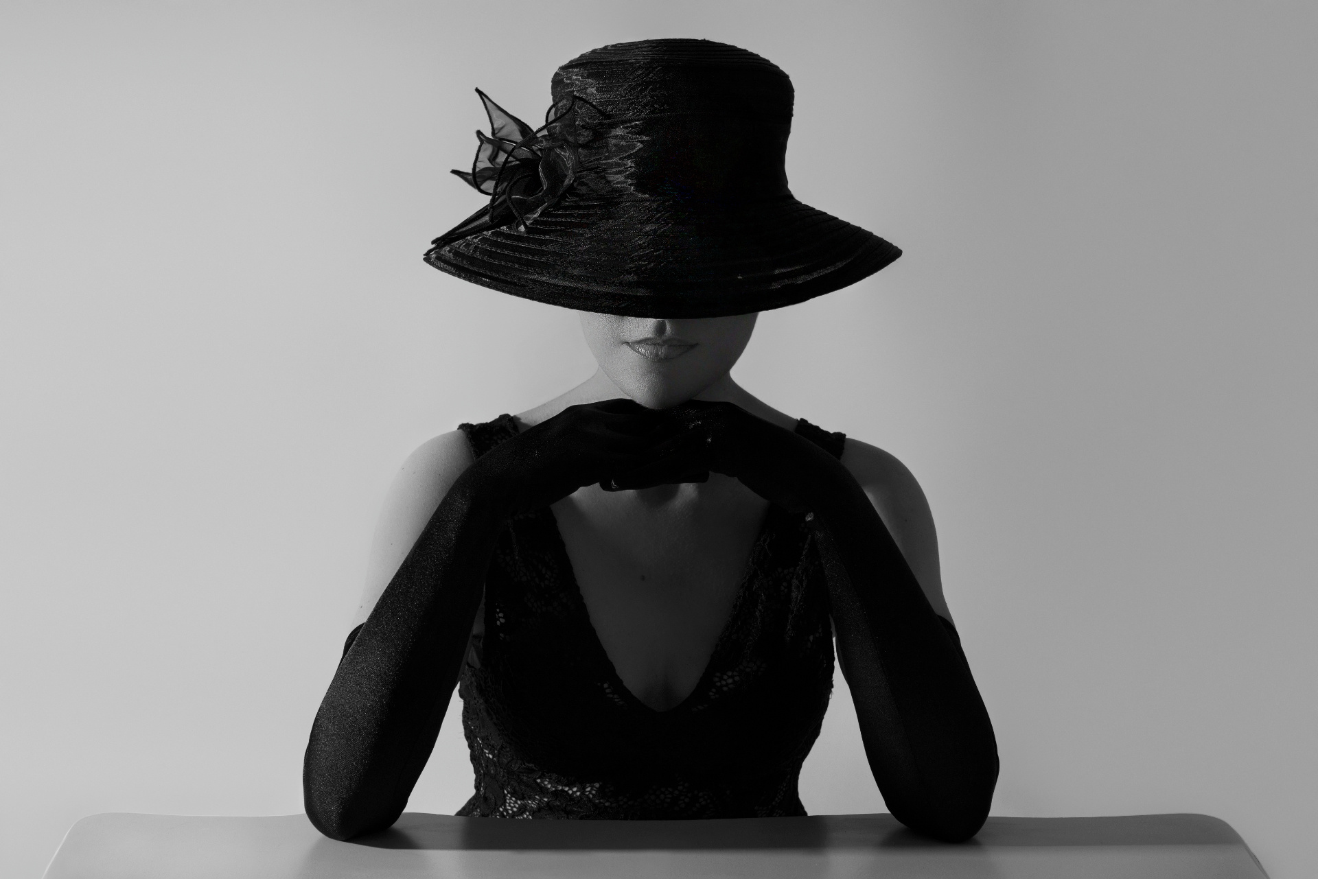 Ritratto fine art in bianco e nero di artista seduta a un tavolo bianco: cappello a tesa larga nero con fiore in organza, abito in pizzo nero scollato sulla schiena e guanti lunghi neri — volto volutamente celato dall'ombra del cappello per un'atmosfera misteriosa ed elegante; book fotografico per artisti di Claudia Spaziani, Castel Guelfo di Bologna