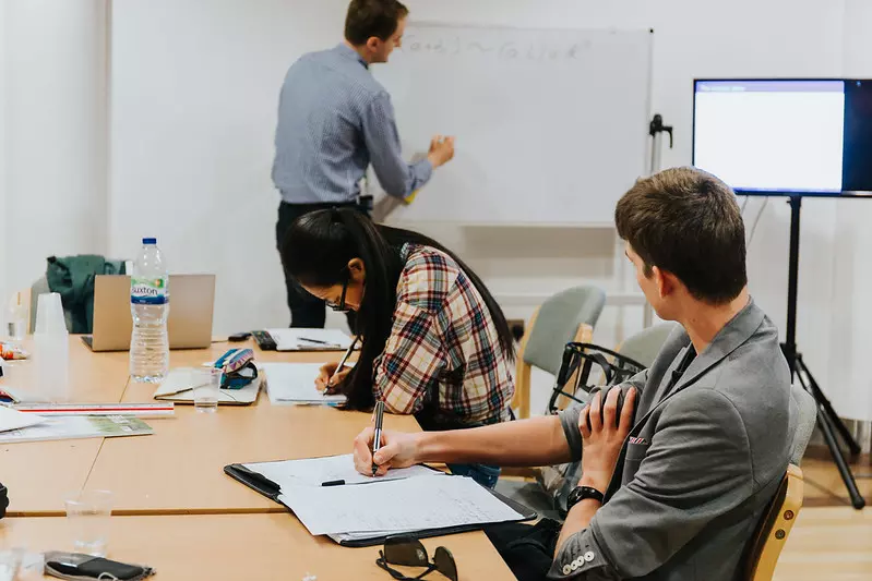 Students in a physics class, observing a tutor writing on a whiteboard