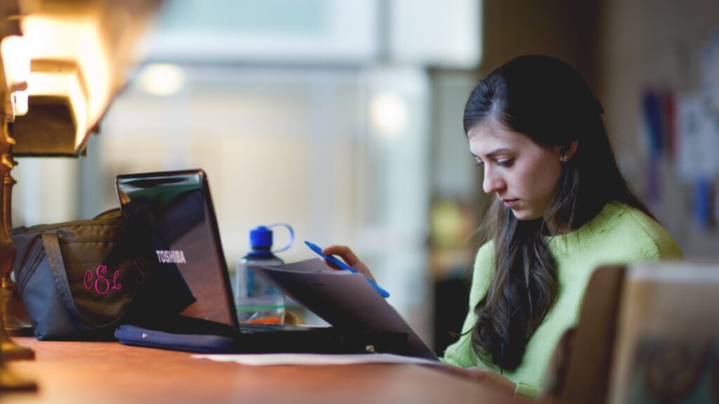 A student attending an online class on their laptop and taking notes using a blue pen