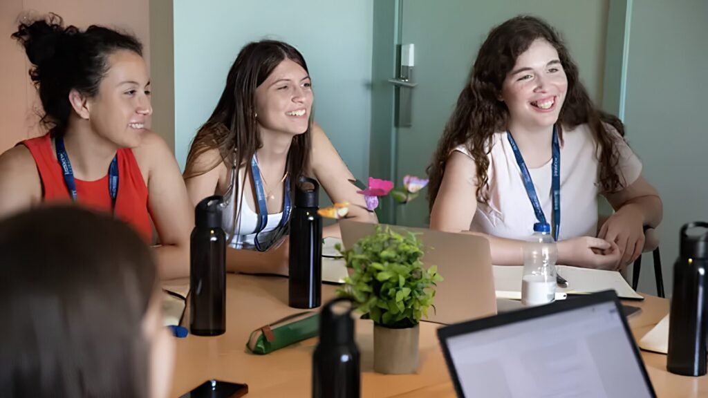 A group of high school students sitting around a table with laptops and water bottles on it, listening to someone speaking.