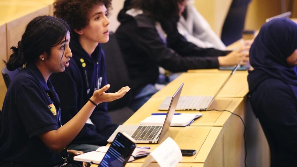 A classroom, focusing on a row of high school students who have devices and notebooks on their tables; a girl, seated, is speaking and gesturing.