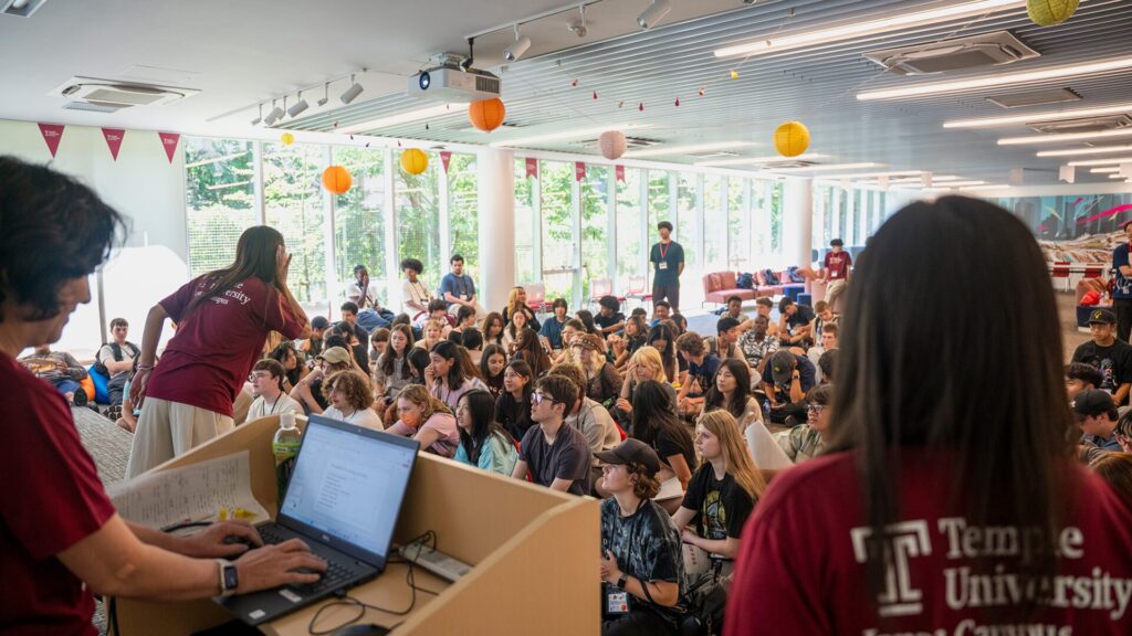 Students seated at a Temple University summer orientation session, with staff in maroon shirts presenting from a podium.