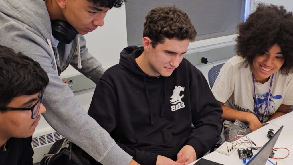 A group of students sitting at the edge of a table and looking at something on a laptop.