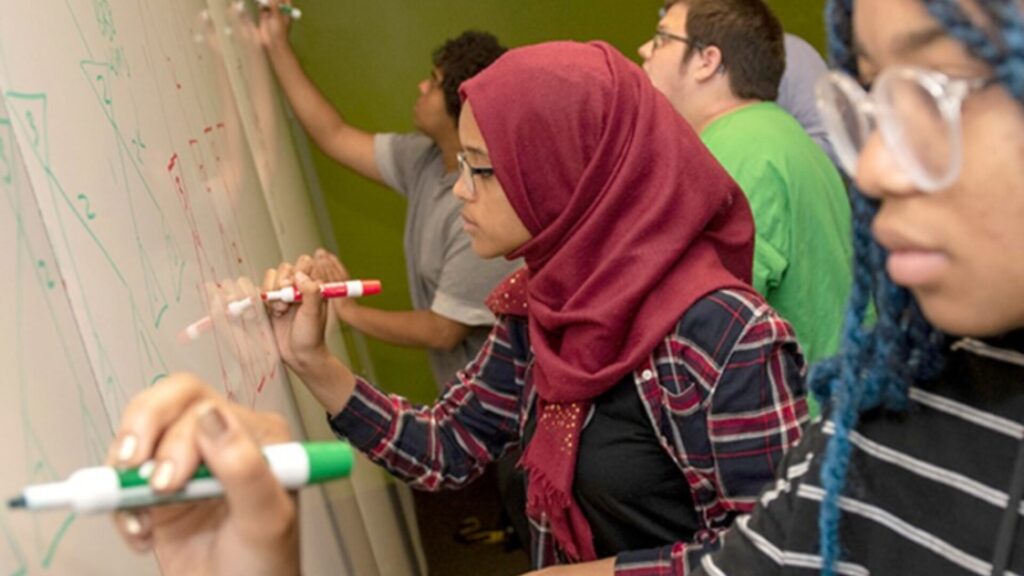 A group of students standing in front of a whiteboard and writing on it with markers