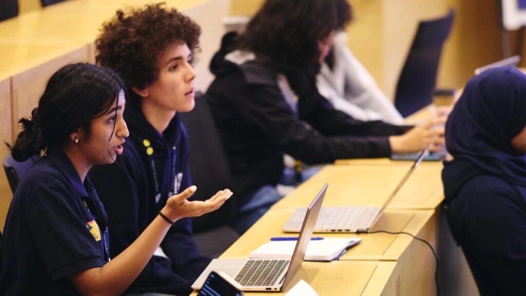 A classroom, focusing on a row of high school students who have devices and notebooks on their tables; a girl, seated, is speaking and gesturing.