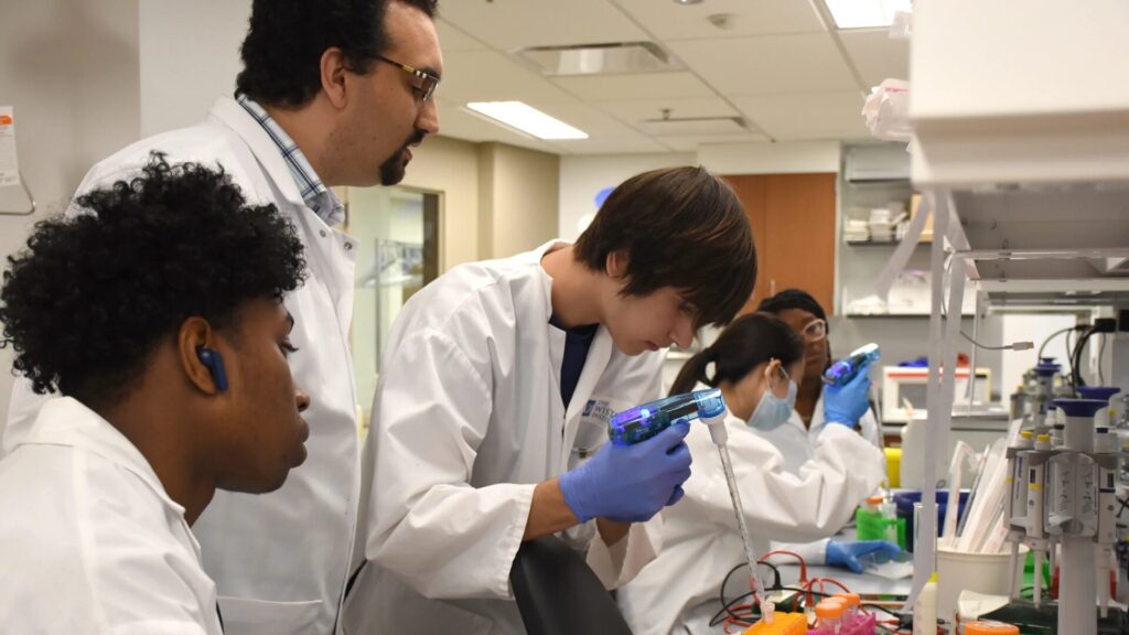 Students wearing lab coats, standing in a lab, and performing experiments as an instructor looks on