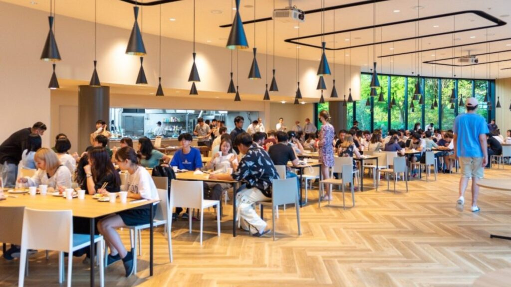 Multiple groups of students are seated at multiple dining tables in a cafeteria, talking to one another.