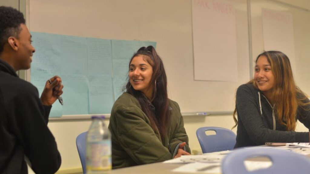 Three young people seated at a classroom table, smiling and chatting during a discussion, with handwritten notes and posters taped to a whiteboard behind them.
