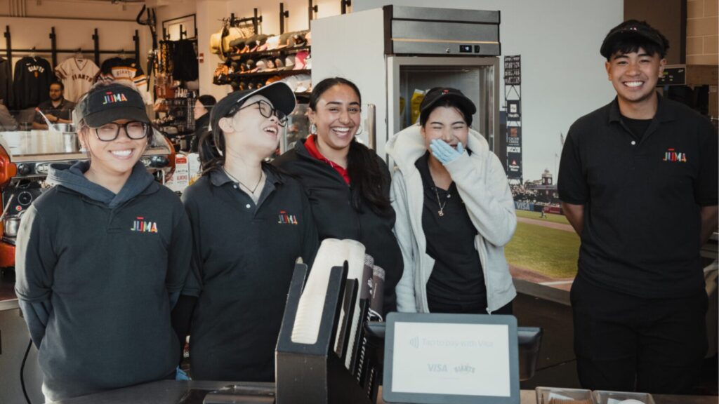 Five students in dark polo shirts and hoodies laughing together behind a merchandise counter with a payment terminal, napkin dispenser, and Giants-branded Visa signage on the counter and jerseys displayed in the background.