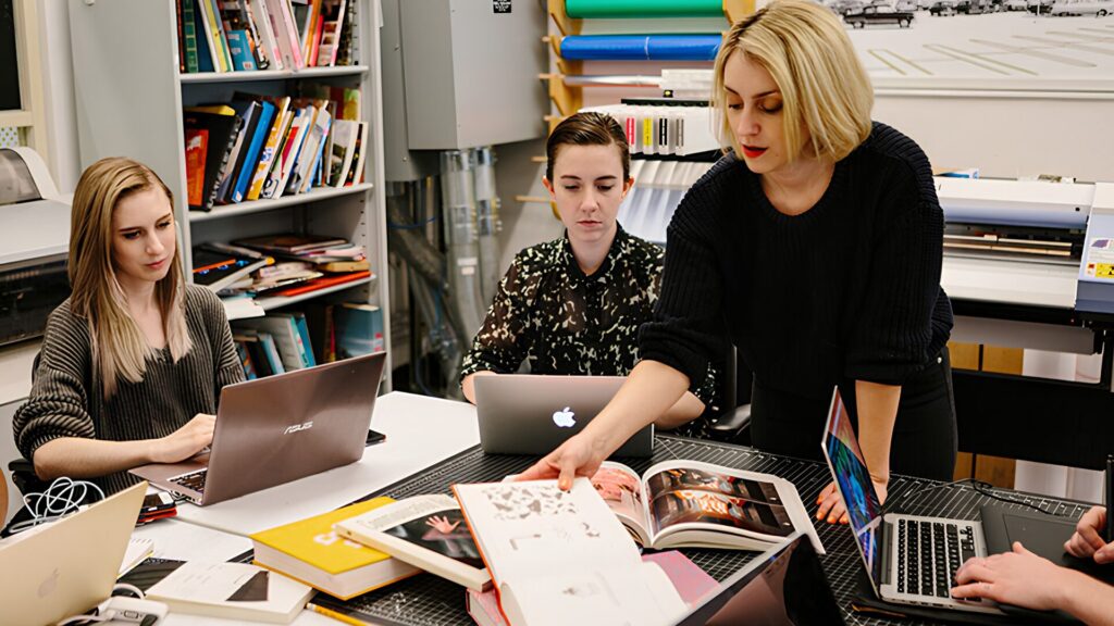 A design instructor showing a book to a table full of seated students with laptops.