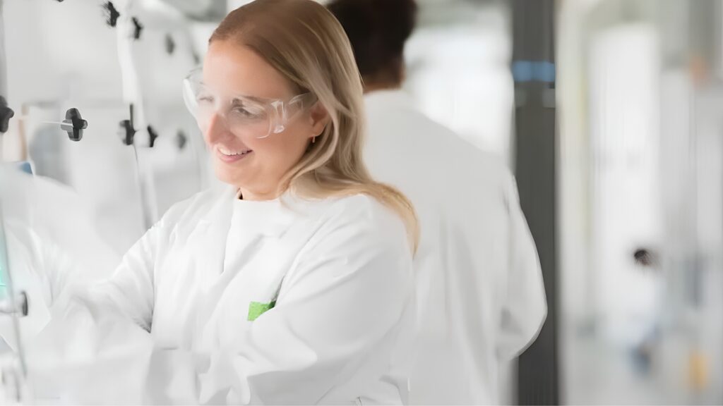 A student wearing a lab coat and green gloves and performing an experiment