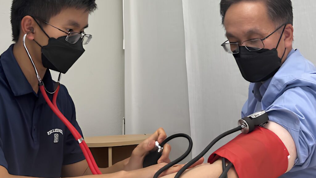 A young man in a stethoscope and mask practices taking blood pressure on an older man's arm with a red cuff.