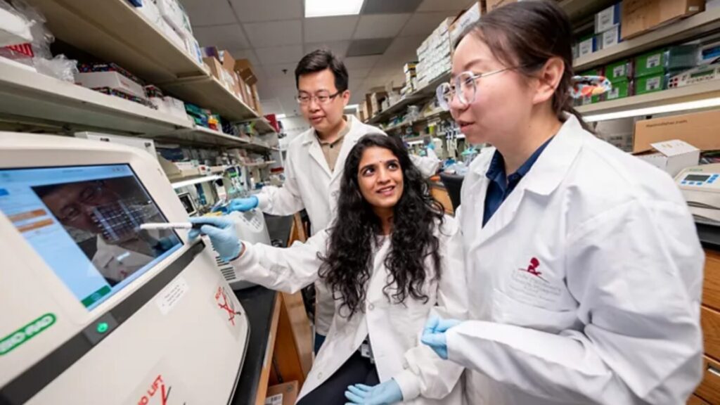 Three lab-coated researchers examine a lab instrument's screen together, one pointing at the display.