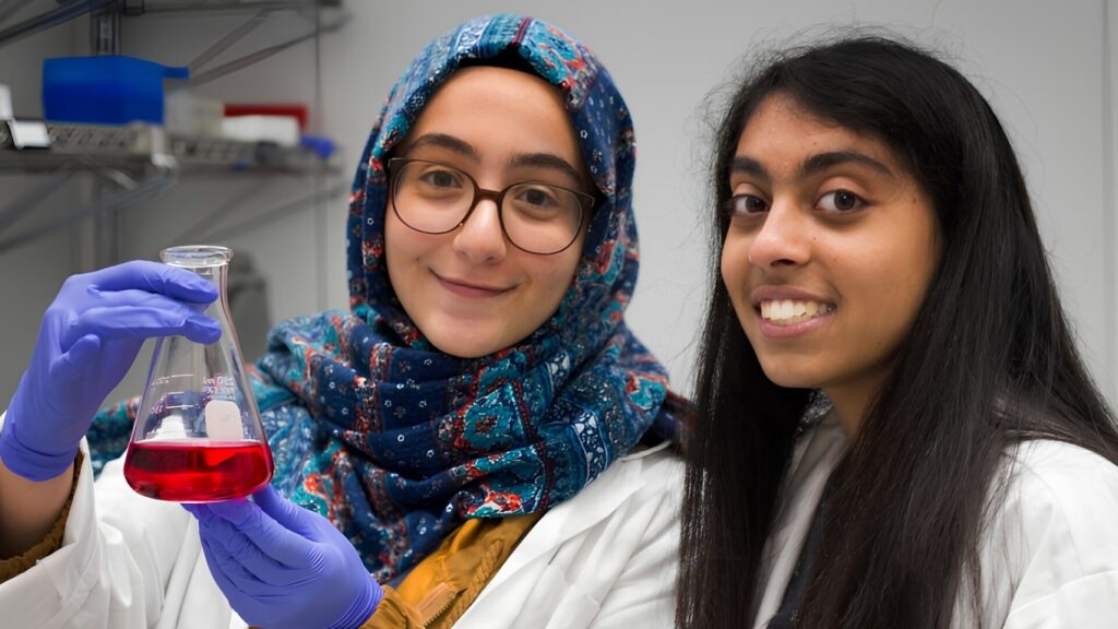 Two students looking into the camera, with one of them holding a beaker of red liquid, and smiling